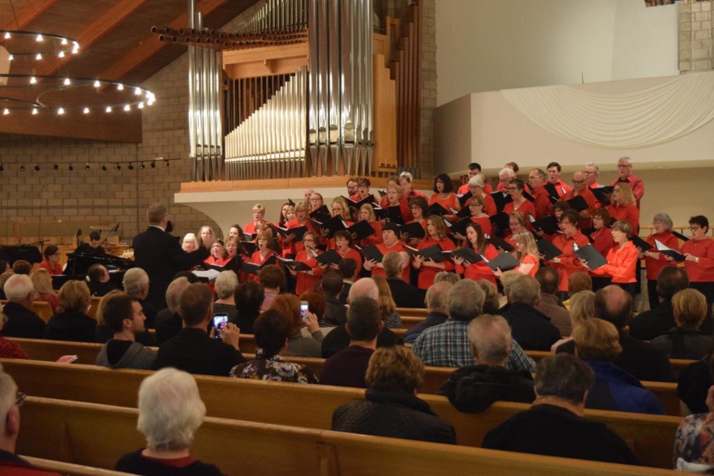 50 singers dressed in red give a concert in a church. A pipe organ can be seen in the background, and the audiences heads can be seen somewhat.